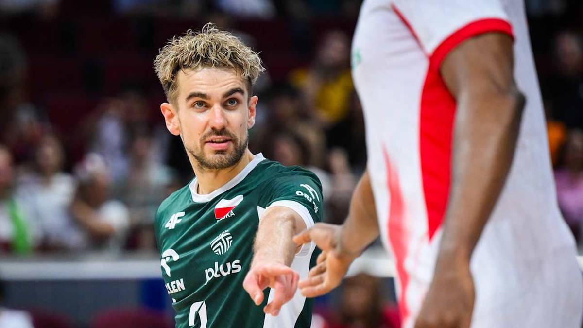 Siatkarz w zielonej koszulce z białymi pasami wskazuje palcem w stronę widza, patrząc na niego z determinacją. Drugi zawodnik w białej koszulce z czerwonymi akcentami stoi bokiem do widza.