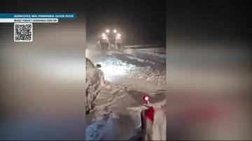 Nocna droga zasypana śniegiem, z widocznymi światłami pojazd&oacute;w i zmagającymi się z warunkami drogowymi ludźmi.