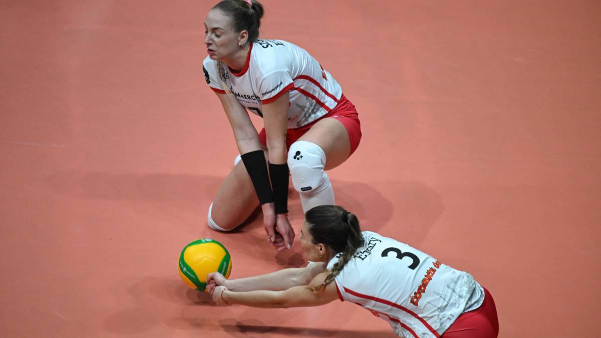 Dwie siatkarki w białych strojach z czerwonymi elementami klęczą na parkiecie, jedna z nich pr&oacute;buje przyjąć piłkę.