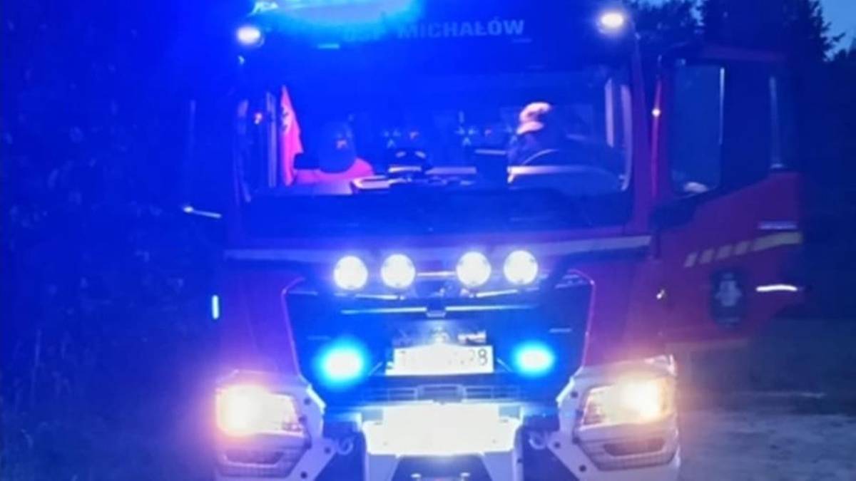 Część przodu wozu strażackiego z włączonymi niebieskimi światłami ostrzegawczymi. Widoczne są reflektory i grill pojazdu.