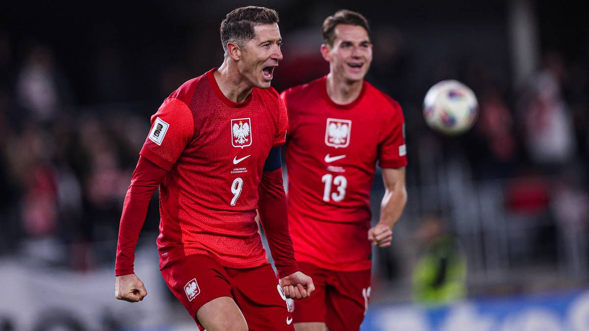 Dwóch polskich piłkarzy w czerwonych strojach, jeden z numerem 9, drugi z numerem 13, świętuje na boisku piłkarskim. Piłkarz z numerem 9 ma otwarte usta w okrzyku radości, a piłkarz z numerem 13 uśmiecha się. W tle widać piłkę i rozmytych kibiców.
