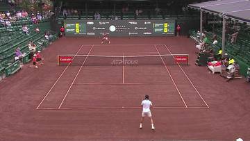 Gracz w białej koszulce i białych spodenkach stoi na ceglanym korcie tenisowym, gotowy do odbicia piłki. Na drugim końcu kortu, w oddali, znajduje się drugi gracz. Widownia siedzi na zielonych trybunach po obu stronach kortu, a nad nimi rozpościera się zadaszenie.