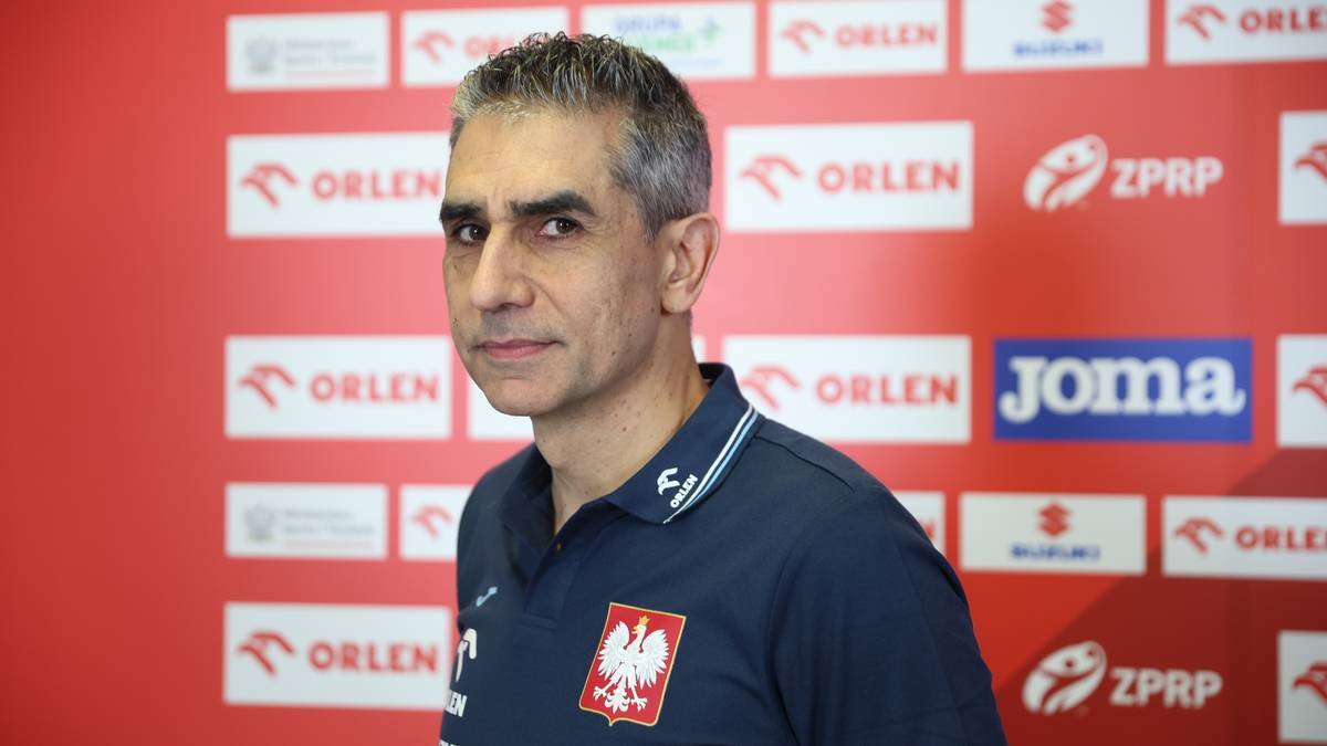 Trener reprezentacji Polski w piłce ręcznej, Jota Gonzalez, stoi przed ścianą z logo sponsor&oacute;w.