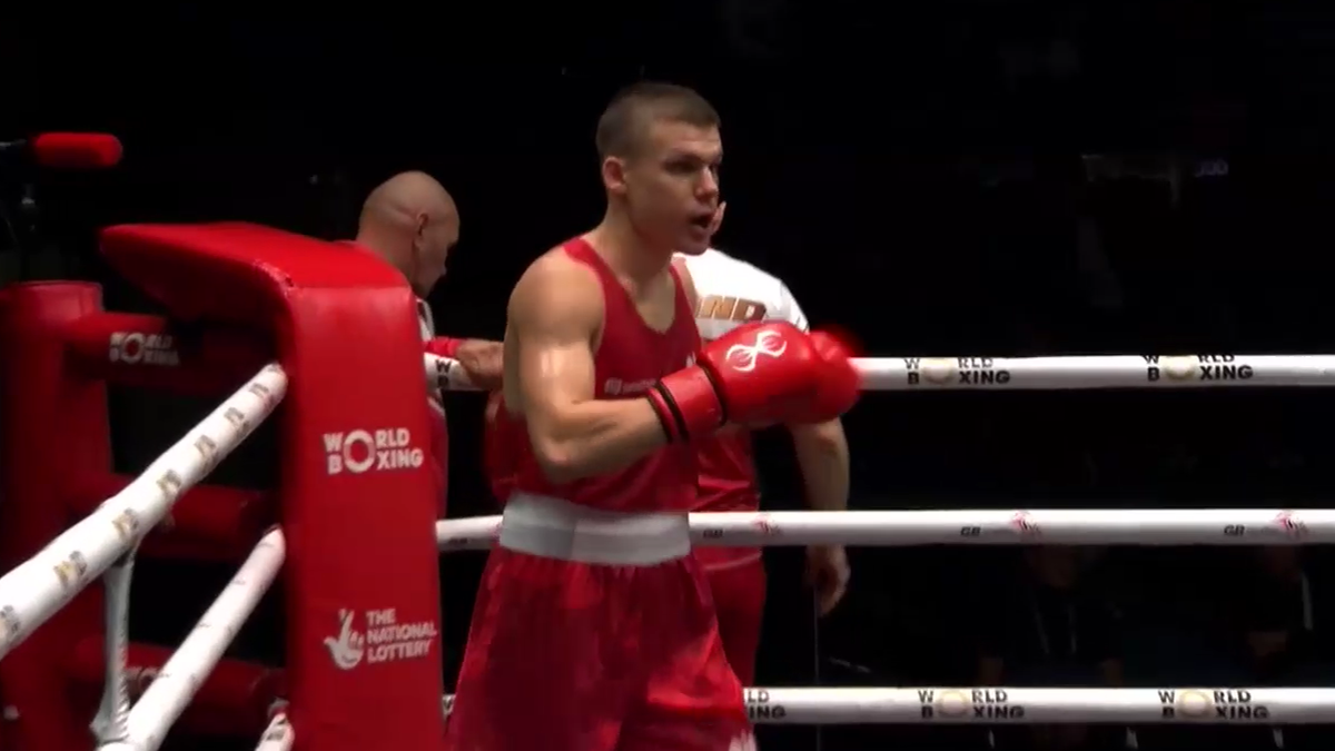 Młody zawodnik boksujący w czerwonym stroju, z czerwoną rękawicą, na tle narożnika i lin ringowych.