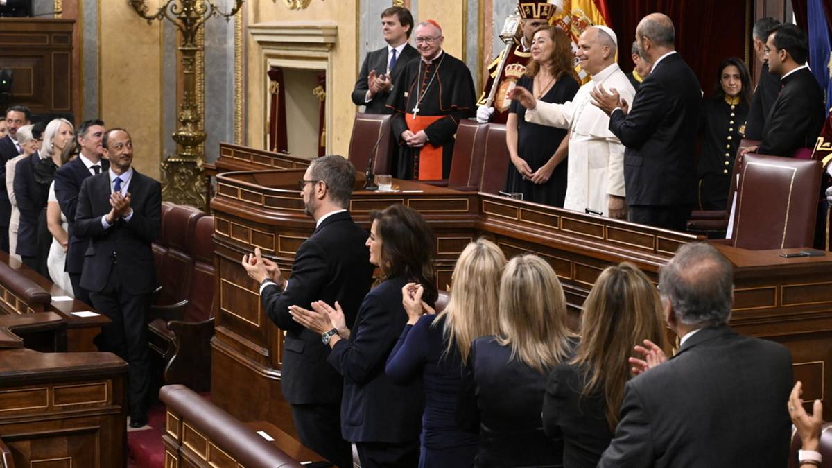 Grupa os&oacute;b w eleganckich strojach, w tym mężczyźni w garniturach i kobiety w sukienkach, stoi w pomieszczeniu przypominającym salę parlamentarną lub uroczystą galerię, część z nich klaszcze, zwracając się w stronę stojącej grupy os&oacute;b na podwyższeniu.