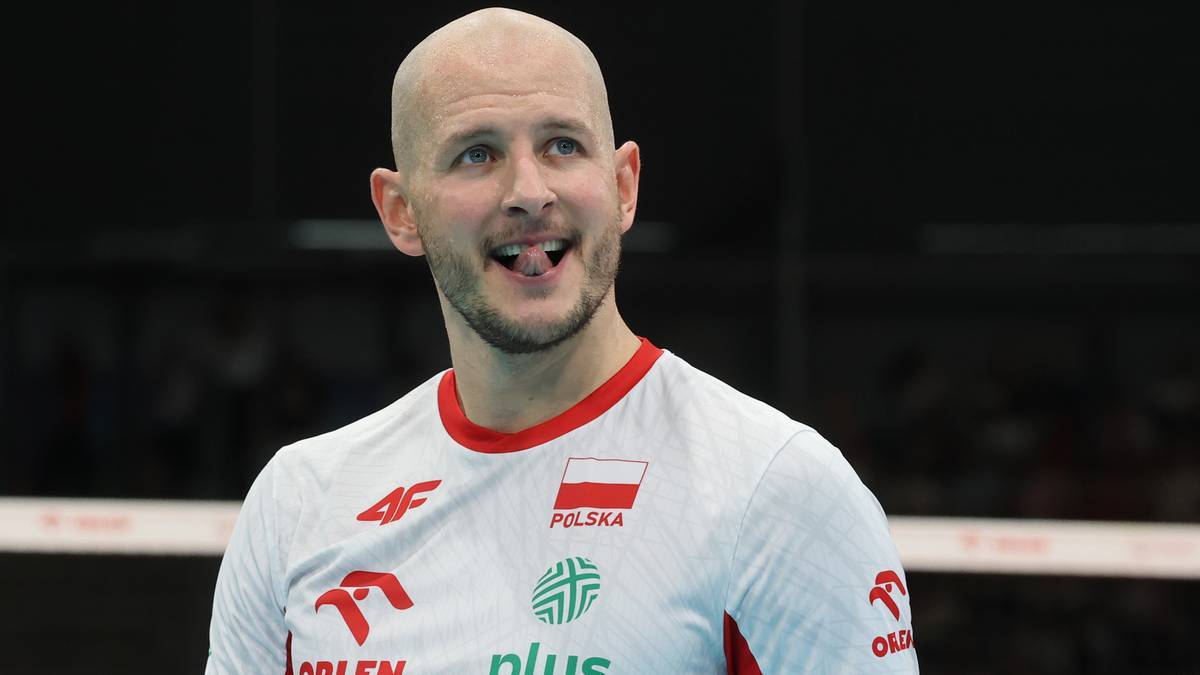 Mężczyzna w białej koszulce sportowej z polską flagą i logo "Polska", z łysą głową i zarostem, uśmiecha się, wystawiając język.