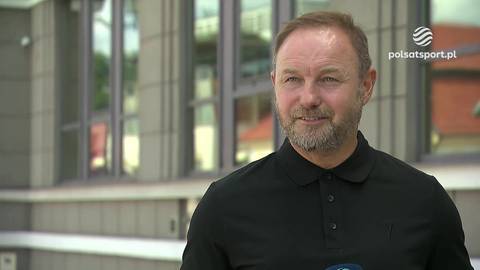 Mężczyzna w średnim wieku z kr&oacute;tkimi, jasnobrązowymi włosami i kilkudniowym zarostem, ubrany w czarną koszulkę polo, uśmiecha się lekko do kamery. W tle widoczny jest budynek z nowoczesną, szarą fasadą i dużymi, prostokątnymi oknami.
