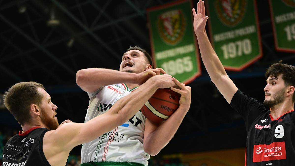 Koszykarz w białej koszulce próbuje rzucić piłkę do kosza, broniony przez dwóch przeciwników w czarnych koszulkach.