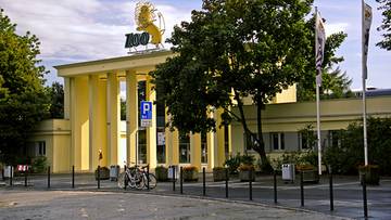 Budynek ZOO Wrocław z napisem "ZOO" na dachu, drzewa i parking przed wejściem.