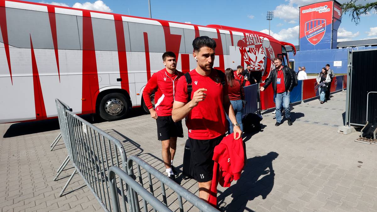 Dwóch piłkarzy w czerwonych koszulkach wychodzi z autokaru, jeden niesie na ramieniu czerwony strój.