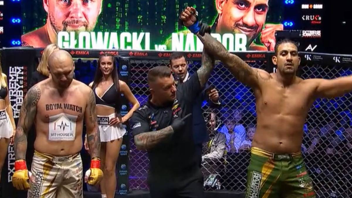 Dwóch mężczyzn w strojach sportowych stoi na ringu, jeden z nich podnosi rękę w geście zwycięstwa, podczas gdy drugi stoi obok niego z opuszczoną ręką. W tle widać publiczność i ekrany.