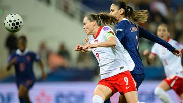 Dwie zawodniczki w białych strojach z czerwonymi detalami ścigają się z piłką nożną, podczas gdy w tle widać inne piłkarki.