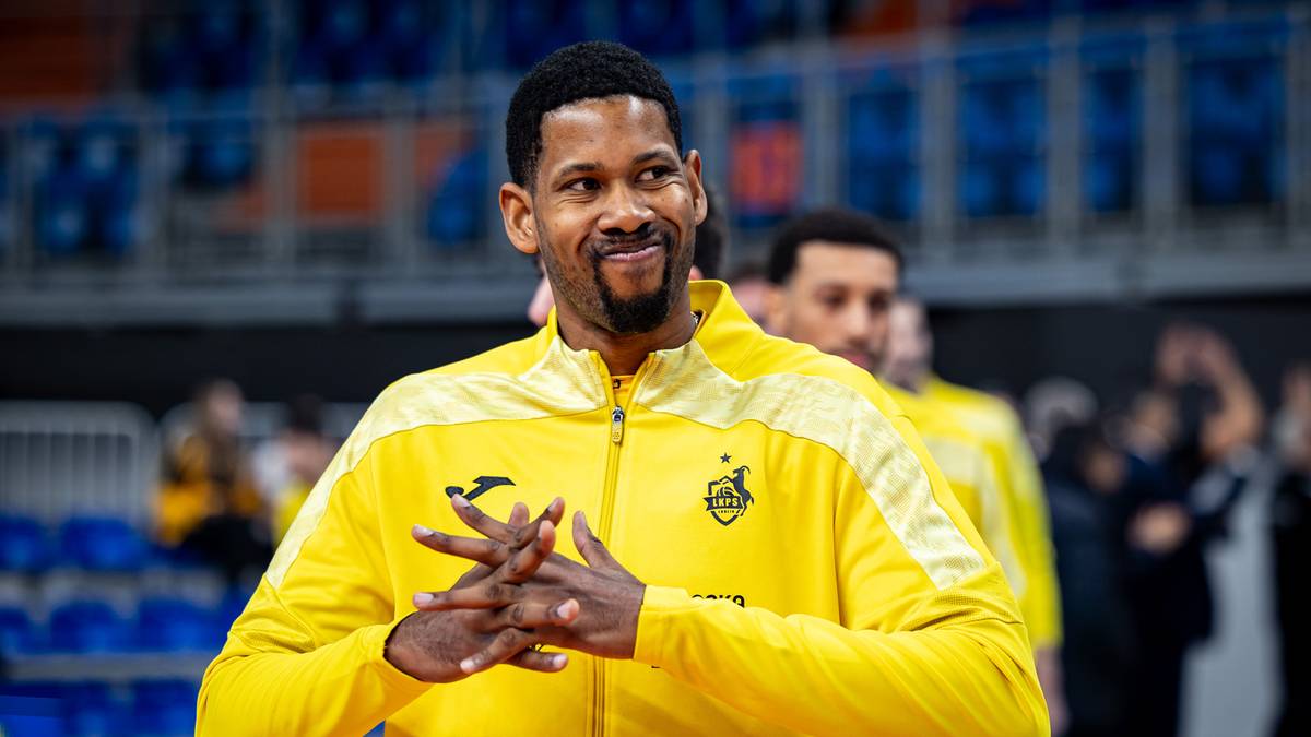 Siatkarz w ż&oacute;łtej bluzie z zapięciem na zamek, z dłońmi splecionymi przed klatką piersiową, uśmiecha się.