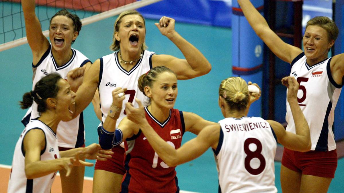 Pięć kobiet w białych i czerwonych strojach sportowych z numerami na koszulkach, świętuje zwycięstwo podczas meczu siatkówki, wznosząc ręce w górę z radości.