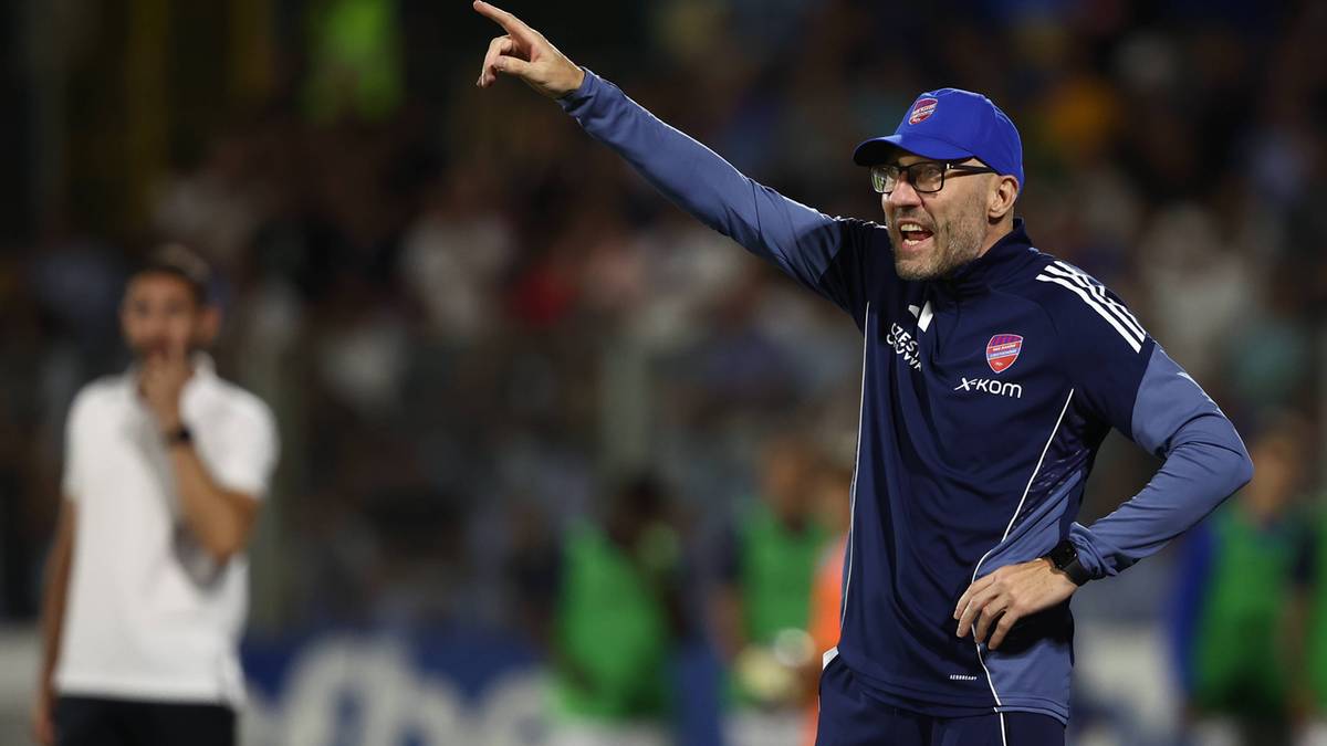 Trener piłkarski w okularach i czapce z daszkiem wskazuje palcem w górę, stojąc na boisku piłkarskim.