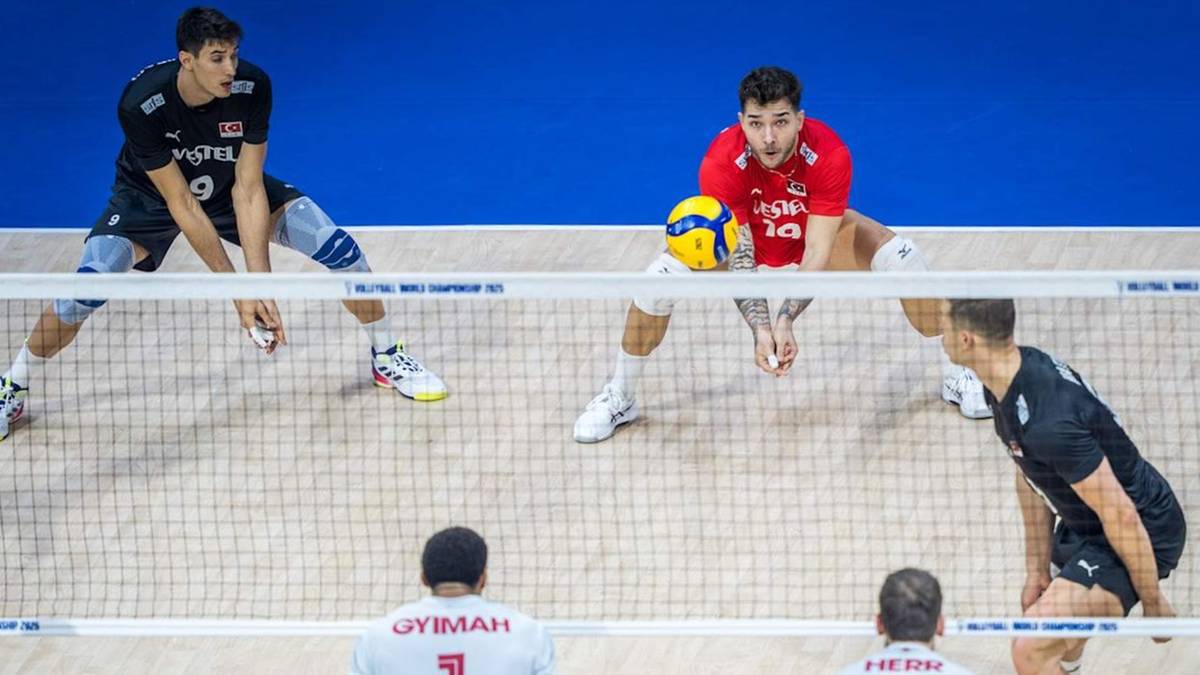 Siatkarze podczas meczu, jeden z nich serwuje piłkę, pozostali są w gotowości do odbioru lub ataku.