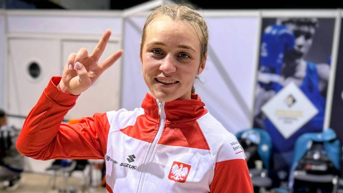 Uśmiechnięta młoda kobieta, Julia Szeremeta, wykonuje znak pokoju palcami wskazującym i środkowym, ubrana w strój sportowy z polskimi barwami narodowymi.