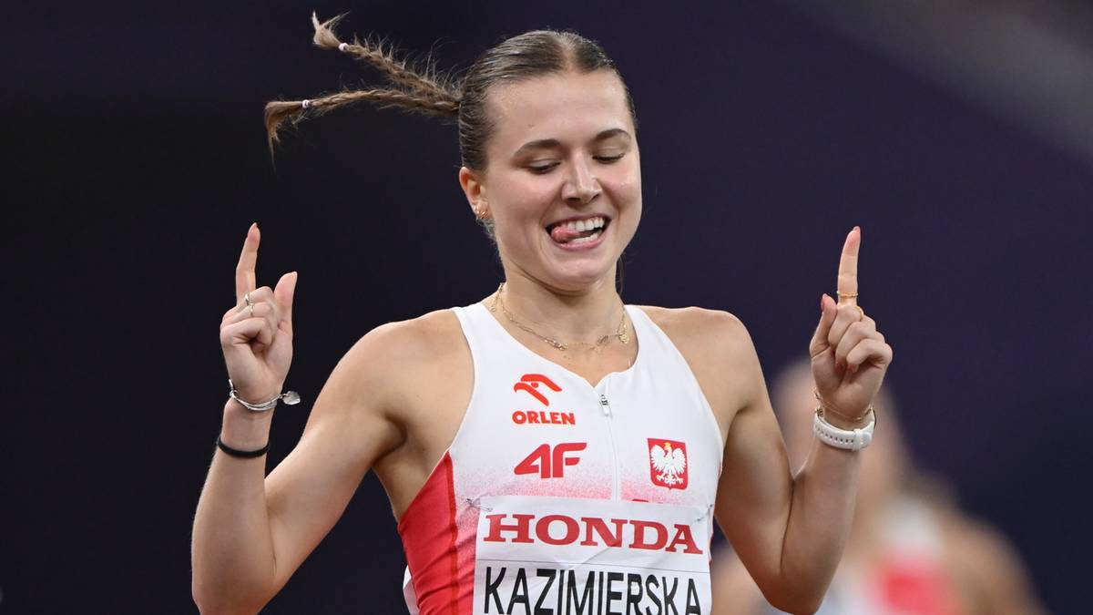 Kobieta lekkoatletka w stroju sportowym z numerem "KAZIMIERSKA" na koszulce, radośnie pokazuje dwa palce w górę.