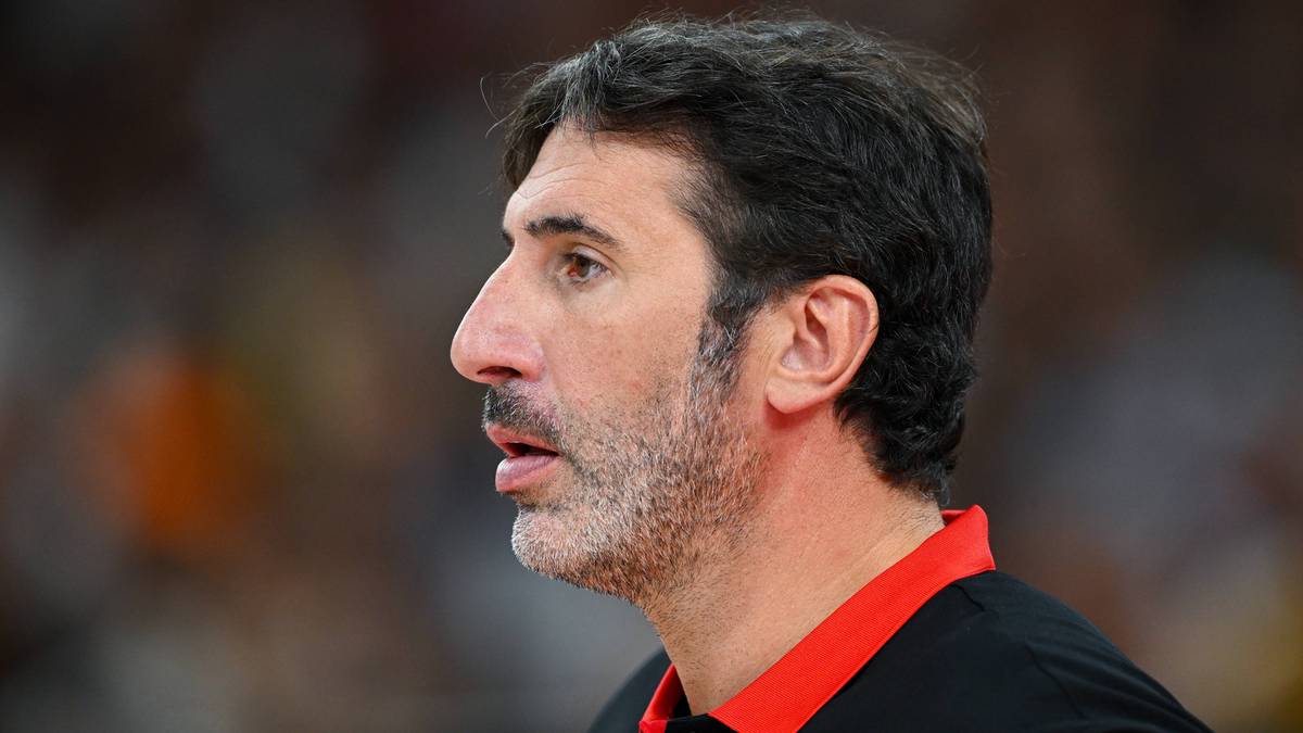 Portret mężczyzny w średnim wieku, patrzącego w bok. Ma krótko przystrzyżone, ciemne włosy i kilkudniowy zarost. Ubrany jest w czarną koszulkę z czerwonym kołnierzykiem.