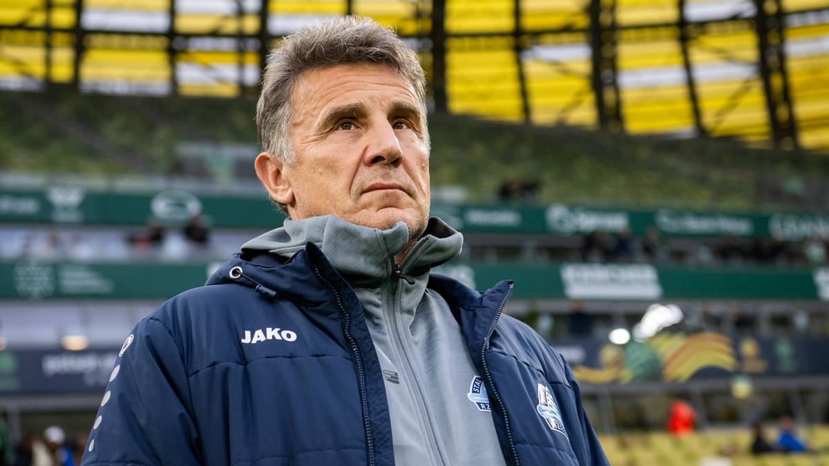 Mężczyzna w średnim wieku, ubrany w niebieską kurtkę sportową z widocznym logo "Jako", patrzy w bok z zamyśloną miną. Tło stanowią trybuny stadionu piłkarskiego.