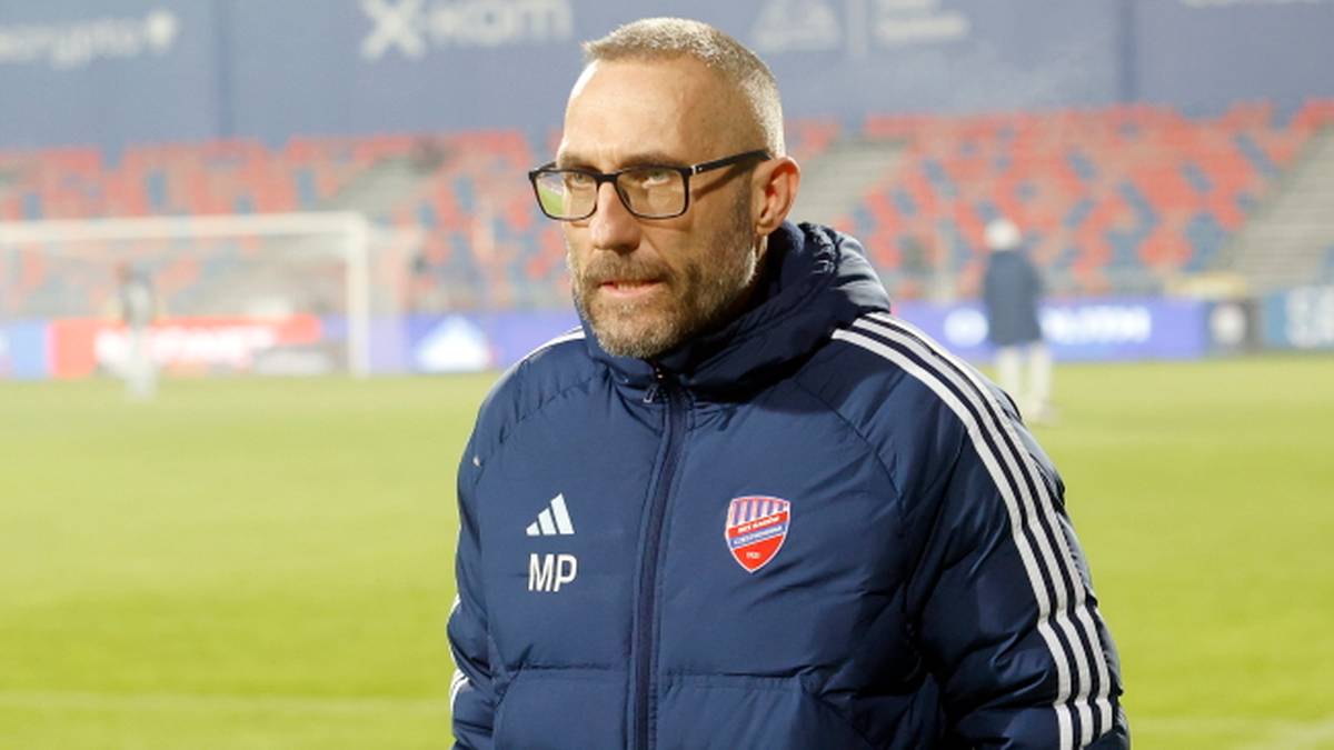 Mężczyzna w okularach i kurtce z logo klubu sportowego stoi na boisku.