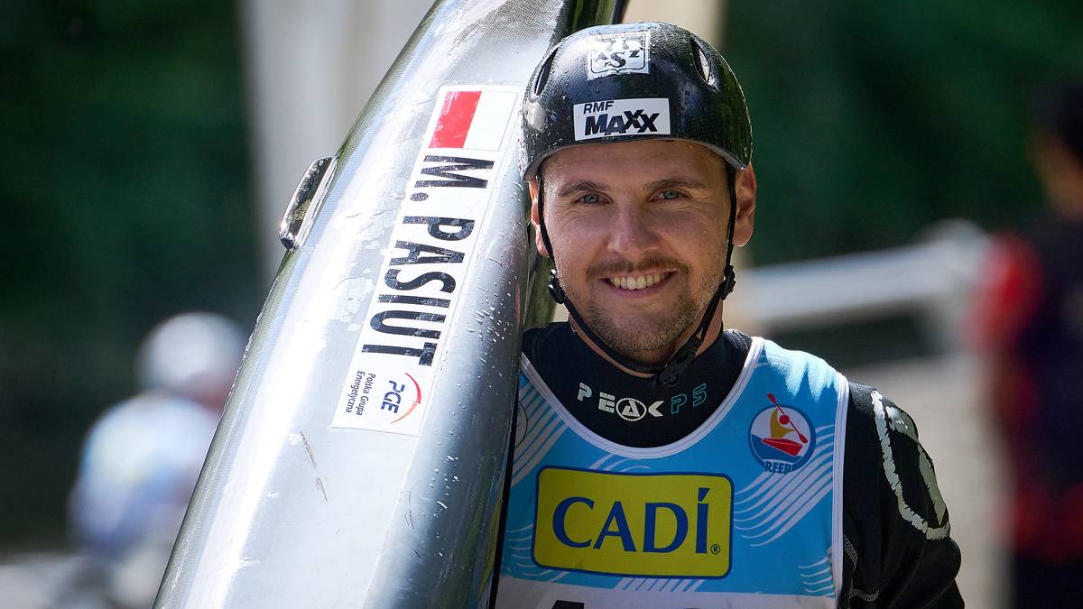 Mężczyzna w kasku i stroju sportowym z napisem "M. PASIUT" na kajaku, uśmiecha się.