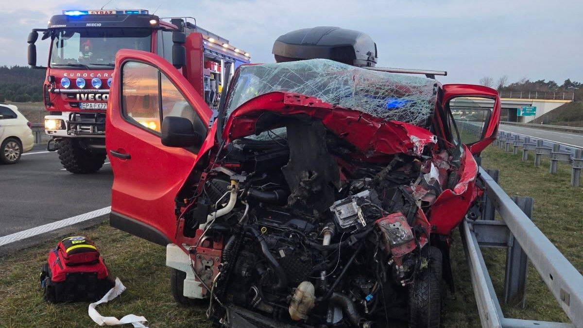 Zniszczony czerwony samochód osobowy po wypadku drogowym, obok wóz strażacki.