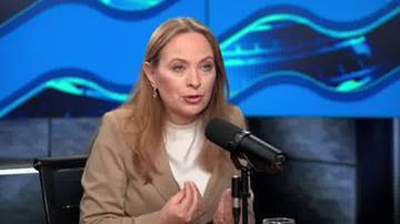 Kobieta w beżowej marynarce siedzi przy stole i m&oacute;wi do mikrofonu.