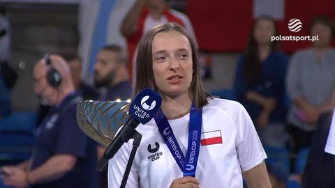 Iga Świątek w białej koszulce z medalem United Cup na szyi, trzymająca mikrofon i puchar.