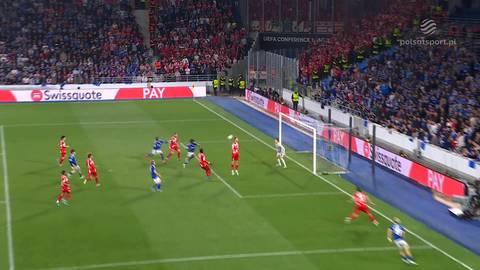 Piłkarze w czerwonych i niebieskich strojach rywalizują o piłkę w polu karnym podczas meczu piłki nożnej.