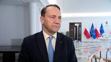 Mężczyzna w średnim wieku, ubrany w ciemny garnitur, jasną koszulę i niebieski krawat, patrzy przed siebie. W tle widoczne są polskie flagi.