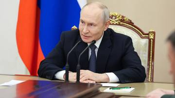 Władimir Putin siedzi przy stole, m&oacute;wiąc do mikrofon&oacute;w. Za nim widoczna jest flaga Rosji.