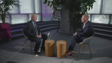 Dw&oacute;ch mężczyzn w garniturach siedzi naprzeciwko siebie na krzesłach w studiu nagraniowym. Pomiędzy nimi znajdują się dwa ż&oacute;łte pufy. W tle widać rośliny i nowoczesne okna.