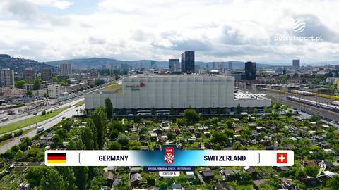 Widok z lotu ptaka na miasto z nowoczesnym stadionem, nad kt&oacute;rym widnieje napis GERMANY i SWITZERLAND z flagami obu kraj&oacute;w, wskazujący na kontekst meczu piłkarskiego.