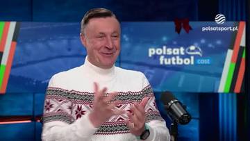 Mężczyzna w białym golfie i świątecznym swetrze z czerwono-czarnym wzorem, uśmiecha się i gestykuluje rękami. Za nim znajduje się ekran z napisem "Polsat Futbol Cast" oraz grafiką stadionu i logo Polsatsport.pl.