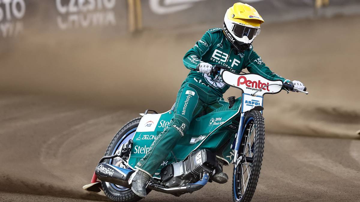 Żużlowiec w zielono-białym kombinezonie i żółtym kasku jedzie na motocyklu po torze żużlowym.