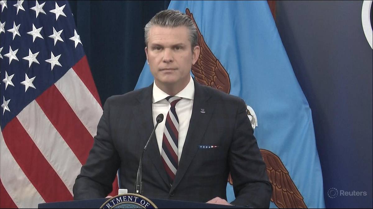 Mężczyzna w garniturze z krawatem stoi przed mikrofonem, za nim widnieją flagi Stan&oacute;w Zjednoczonych i niebieska flaga z symbolem orła.