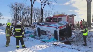 Rozbita karetka pogotowia leżąca na boku na poboczu drogi, w pobliżu stoją ratownicy w kamizelkach i strażacy w mundurach.