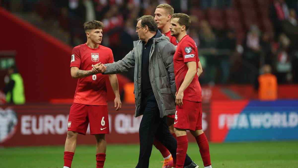 Trener polskiej reprezentacji piłkarskiej, ubrany w szary płaszcz, wita się z trzema zawodnikami w czerwonych strojach piłkarskich na zielonej murawie stadionu.