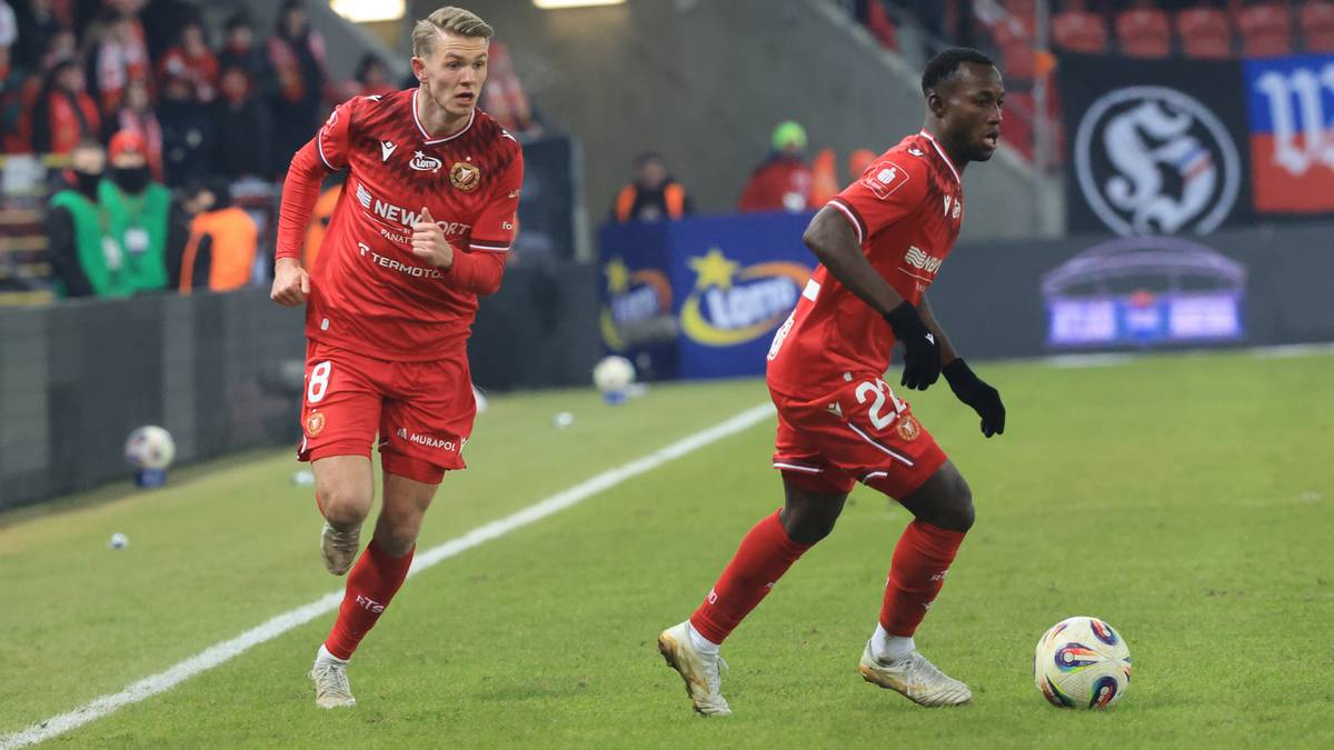 Dw&oacute;ch piłkarzy w czerwonych koszulkach, jeden biegnie z piłką, drugi za nim, na boisku piłkarskim.