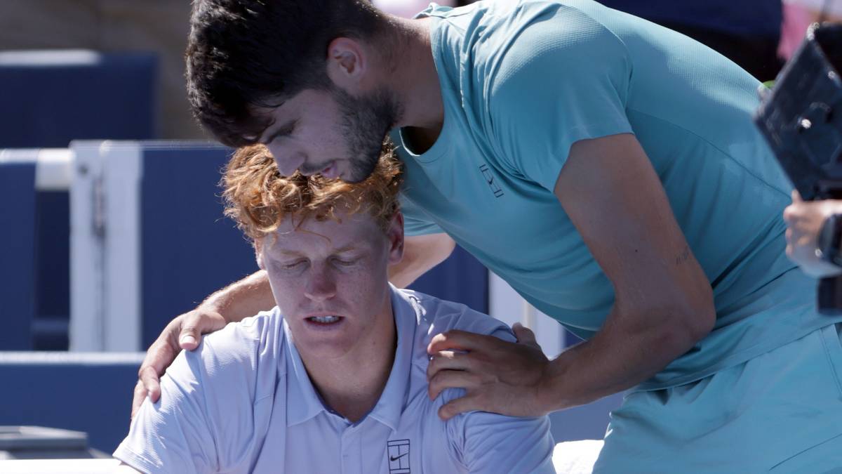 Dw&oacute;ch tenisist&oacute;w na korcie tenisowym. Jeden opiera głowę na drugim, prawdopodobnie z powodu kontuzji lub zmęczenia. Obaj są ubrani w sportowe stroje.