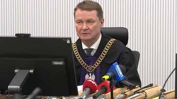 Sędzia w todze sądowej z łańcuchem na szyi siedzi za biurkiem przed komputerem, otoczony mikrofonami.