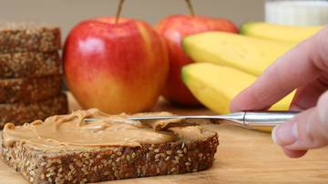 Ktoś rozsmarowuje masło orzechowe na kromce ciemnego chleba razowego. W tle widać jabłka i banany.