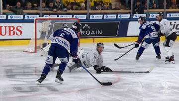 Gracze w hokeju na lodzie rywalizują o krążek w pobliżu bramki. Zawodnik w białym stroju leży na lodzie, próbując odebrać krążek graczowi w niebieskim stroju.