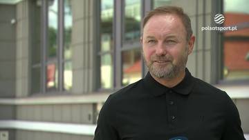 Mężczyzna w średnim wieku z kr&oacute;tkimi, jasnobrązowymi włosami i kilkudniowym zarostem, ubrany w czarną koszulkę polo, uśmiecha się lekko do kamery. W tle widoczny jest budynek z nowoczesną, szarą fasadą i dużymi, prostokątnymi oknami.