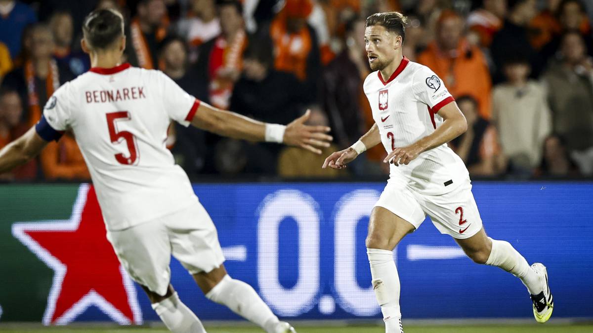 Dw&oacute;ch piłkarzy reprezentacji Polski w białych strojach grających na boisku piłkarskim. Jeden z nich, z numerem 5 na plecach, odwr&oacute;cony plecami do widza, wykonuje gest w kierunku swojego kolegi. Drugi piłkarz, z numerem 2, biegnie w jego stronę. W tle widać kibic&oacute;w na trybunach oraz fragment wyniku sportowego na ekranie.