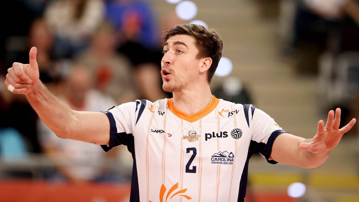 Siatkarz w białej koszulce z numerem 2 i pomarańczowymi wstawkami, wykonuje gest rękami.