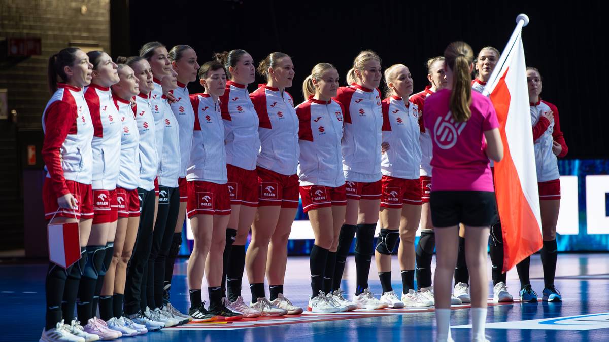 Drużyna szczypiornistek w białych strojach z czerwonymi elementami stoi w szeregu. Jedna z zawodniczek, ubrana w różowy strój, trzyma biało-czerwoną flagę.