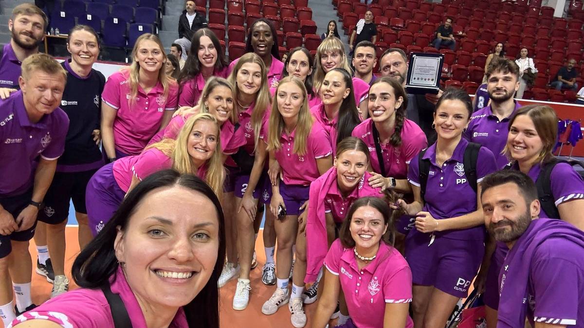 Grupa siatkarzy i siatkarek, ubranych w stroje sportowe w kolorach fioletowym i różowym, pozuje do zdjęcia w hali sportowej.