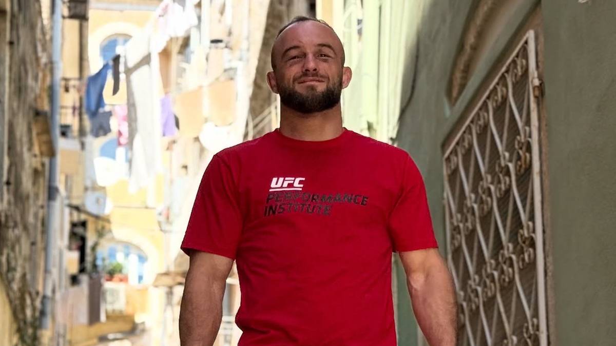 Mężczyzna w czerwonej koszulce UFC z krótkim zarostem uśmiecha się do obiektywu.